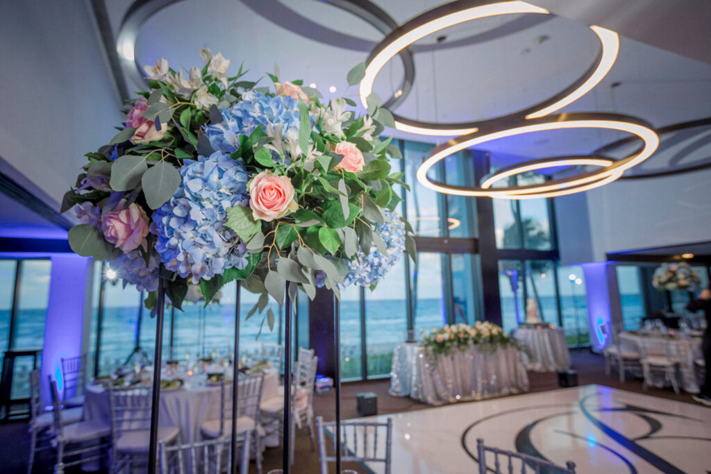 A stunning elevated floral centerpiece featuring lush blue hydrangeas, soft pink roses, white blooms, and elegant greenery. This luxury wedding floral arrangement by Zeta Event Productions enhances the sophisticated ambiance of a West Palm Beach oceanfront wedding. Event planning and designer Roxy Quevedo curated the perfect elegant décor, captured by Victoria Machin Photography.