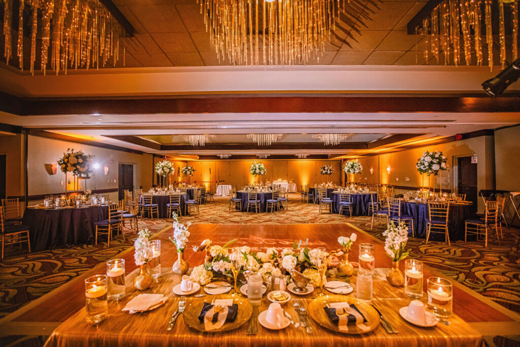 Amber uplighting creates a warm and elegant ambience at a wedding reception ballroom with gold and white décor.