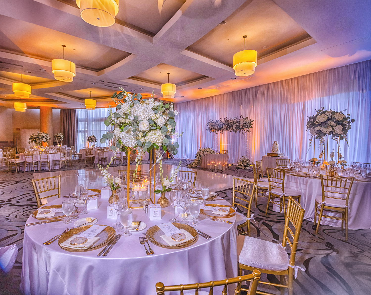 Elegant wedding reception setup with tall white floral centerpieces, gold Chiavari chairs, blush drapery, and ambient lighting by Zeta Event Productions in South Florida.