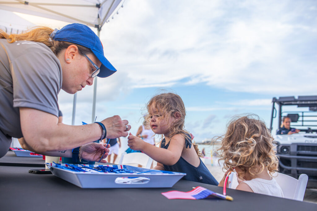 A Zeta Event Productions team member helps young children with patriotic crafts during a 4th of July celebration on the beach, with American flags and a scenic coastal backdrop.