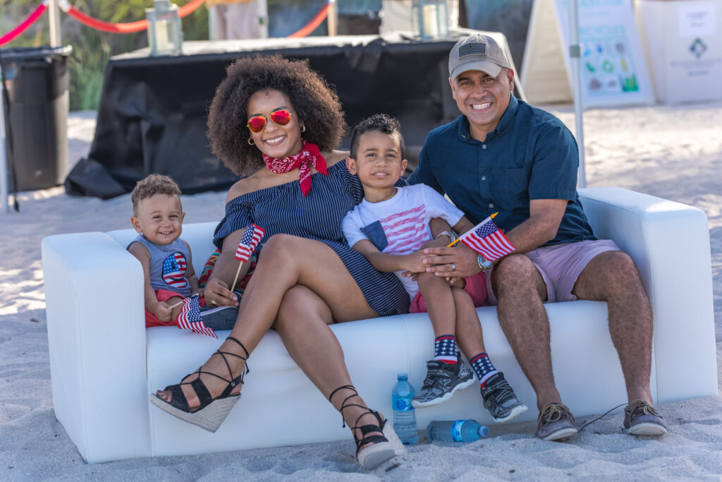 A joyful family of four celebrating the 4th of July on the beach in Bal Harbour, holding American flags and dressed in red, white, and blue, captured by Victoria Machin Photography.